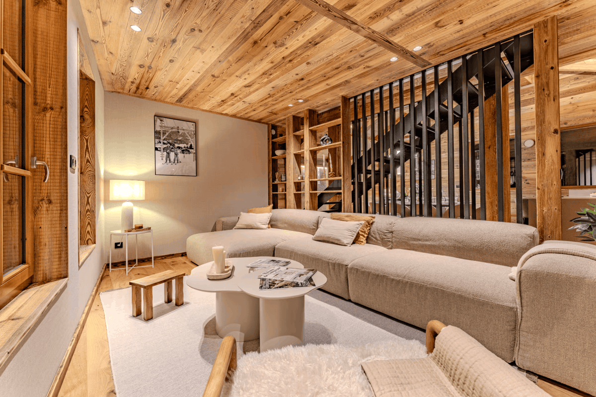A modern U-shaped sectional sofa in light beige fabric, styled in a cozy, rustic chalet living room with a wood-paneled ceiling, a round white coffee table, and a black metal staircase in the background.