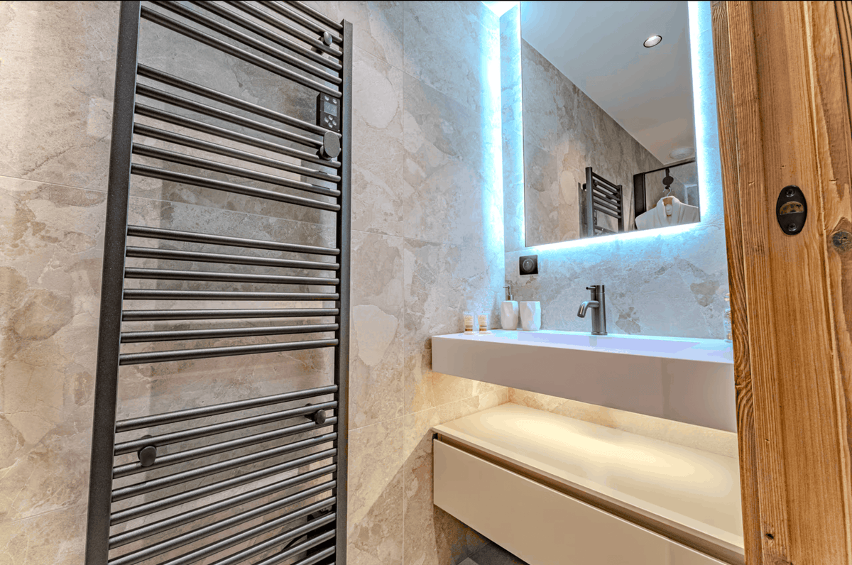A modern bathroom interior featuring a tall dark gray heated towel rail, a white floating vanity, a large backlit mirror, and beige marble walls.