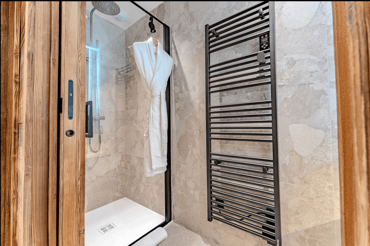 A modern bathroom interior featuring a walk-in shower enclosure with a white robe hanging inside, beige stone walls, and a black heated towel rail.