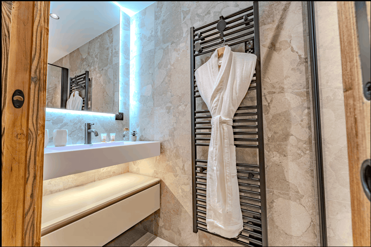 A white bathrobe hangs on a black heated towel rail in a modern bathroom with beige stone walls, a floating white vanity, and blue LED lighting.