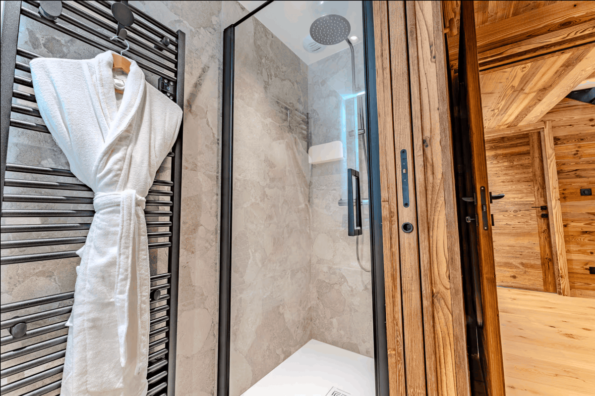 A white plush bathrobe hangs on a black heated towel rail beside a glass shower enclosure in a modern bathroom, with a wood-paneled room visible through an open door.