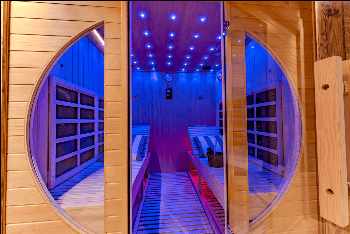 A wooden sauna interior with circular window openings, blue ambient lighting, wooden benches, and a slatted walkway, photographed from outside the window.