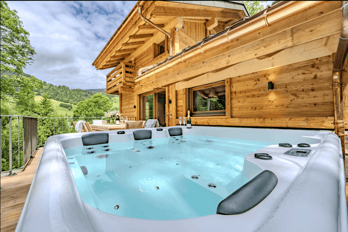 A white outdoor hot tub with light blue water, set on a wooden deck beside a large wood-paneled chalet with a scenic mountain view.