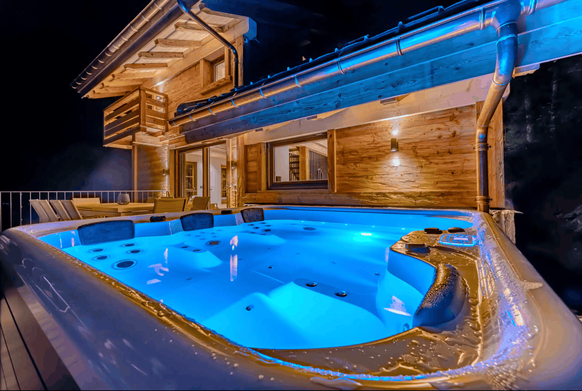 An outdoor hot tub with bright blue illuminated water, set beside a modern wooden chalet at night.