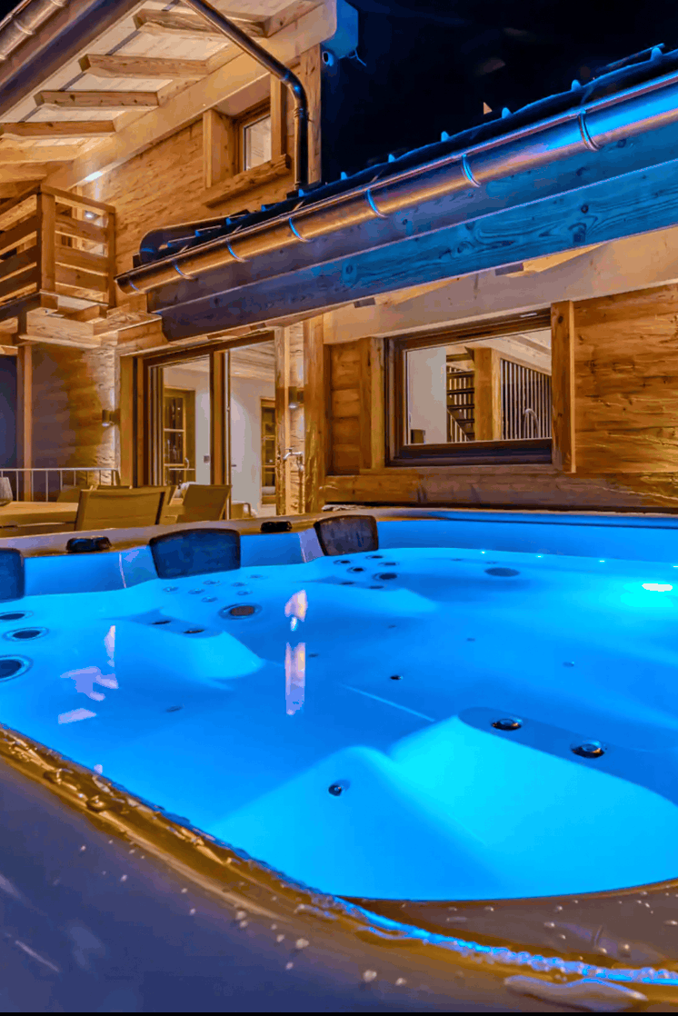 A large outdoor hot tub with bright blue illuminated water, set beside a wooden chalet at night.