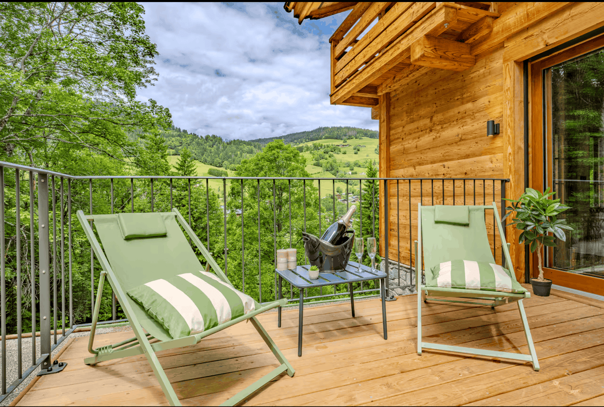 A wooden deck balcony with two green lounge chairs, a small table with an open bottle, and a scenic view of green hills and trees under a cloudy sky.