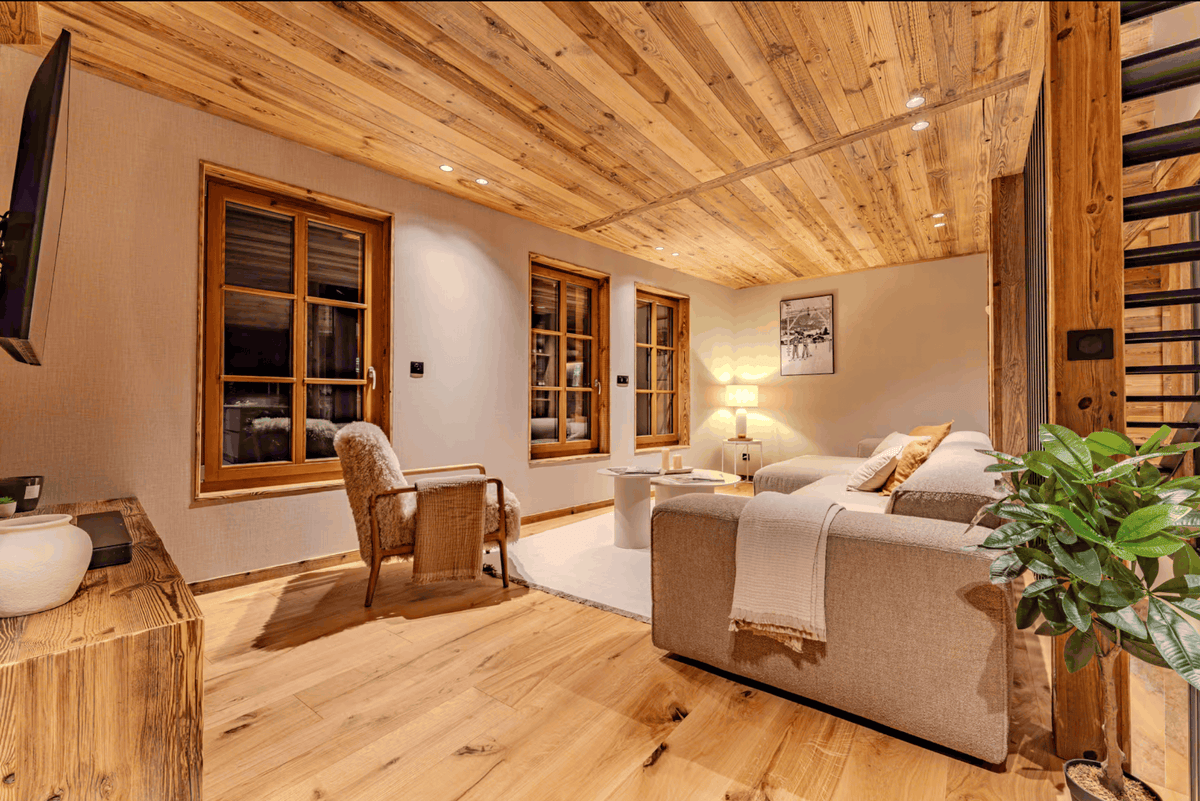 A modern, cozy chalet-style living room with light wood plank ceilings and floors, featuring a beige sofa, a rustic armchair, a potted green plant, and large wood-framed windows.