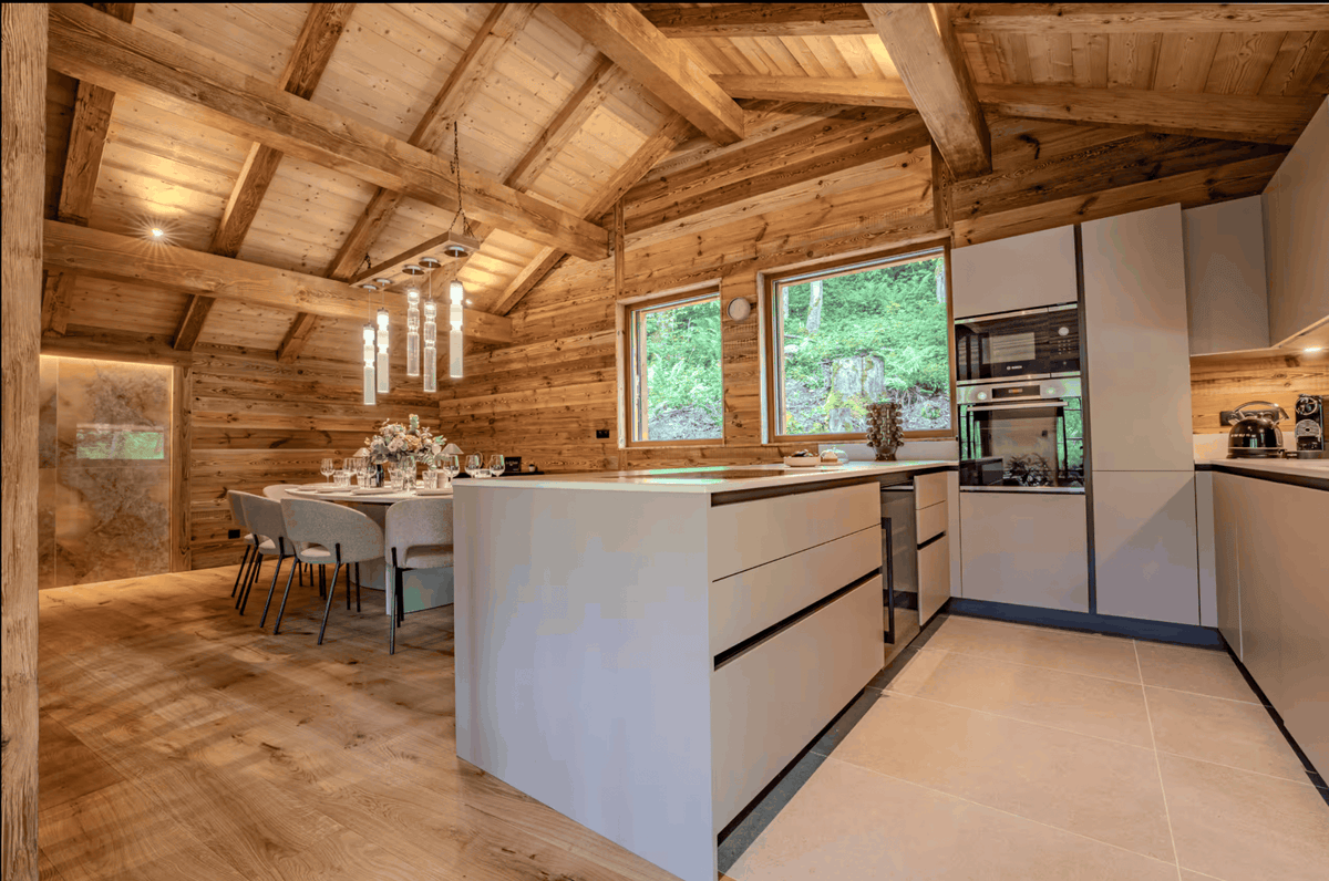 A modern kitchen and dining area in a rustic wooden chalet, featuring white kitchen cabinets, a central island, built-in appliances, and a dining table set under a high wood-beamed ceiling.
