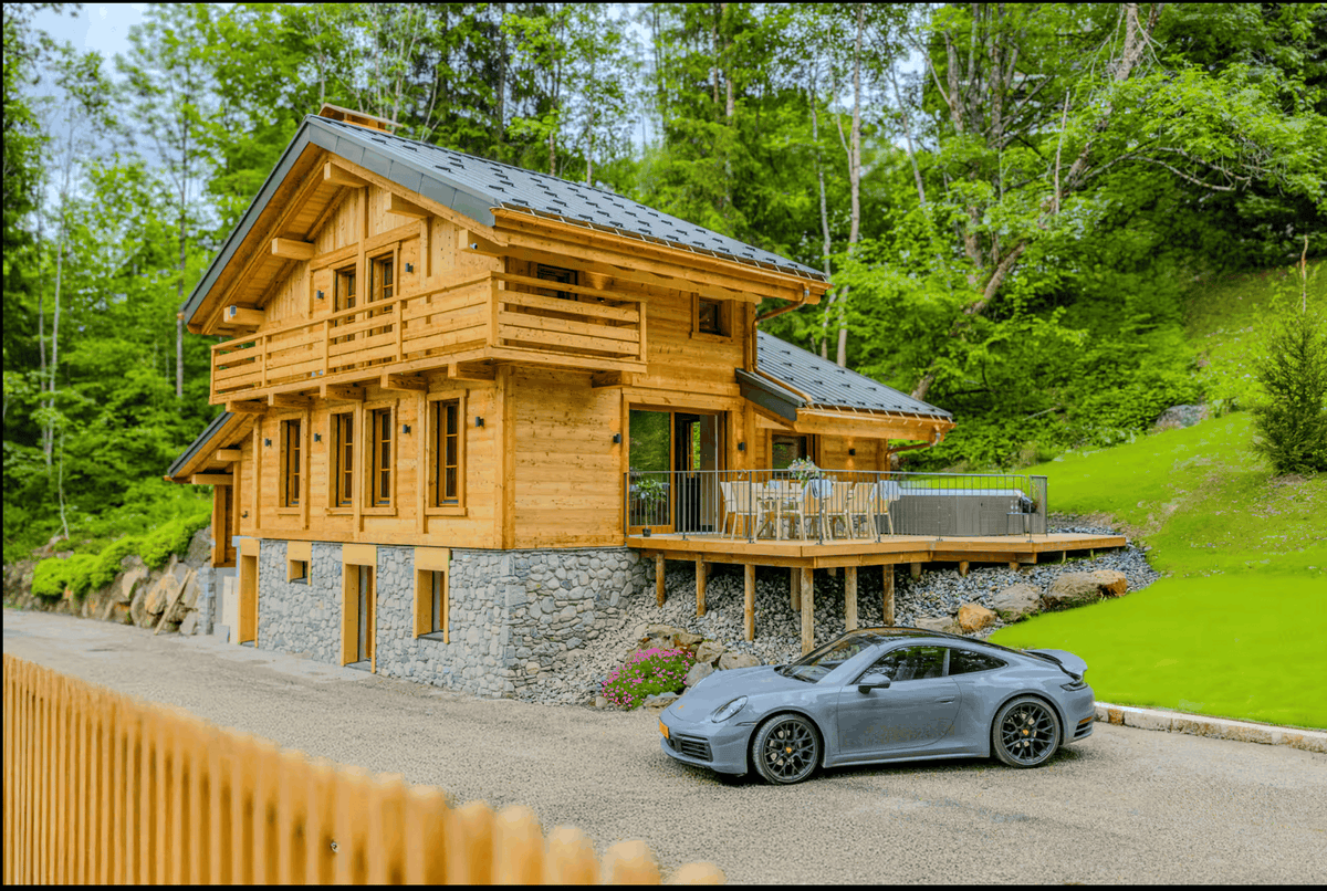 A gray Porsche sports car parked on a gravel driveway in front of a modern wooden chalet with a stone foundation, surrounded by a lush green forest and grassy hillside.