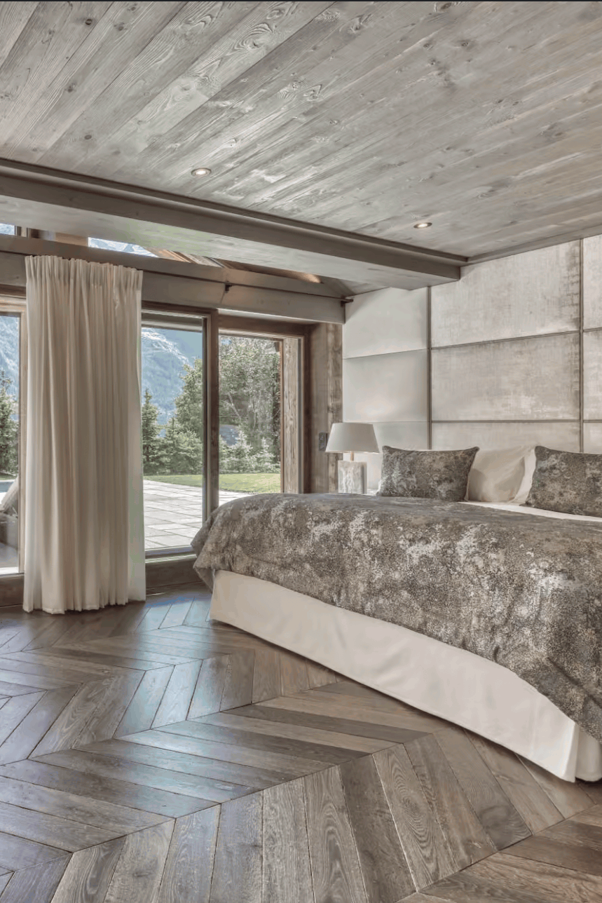 A luxury bedroom interior with a large bed dressed in gray and white patterned bedding, herringbone wood flooring, paneled walls, and floor-to-ceiling windows overlooking a mountain landscape and outdoor patio.