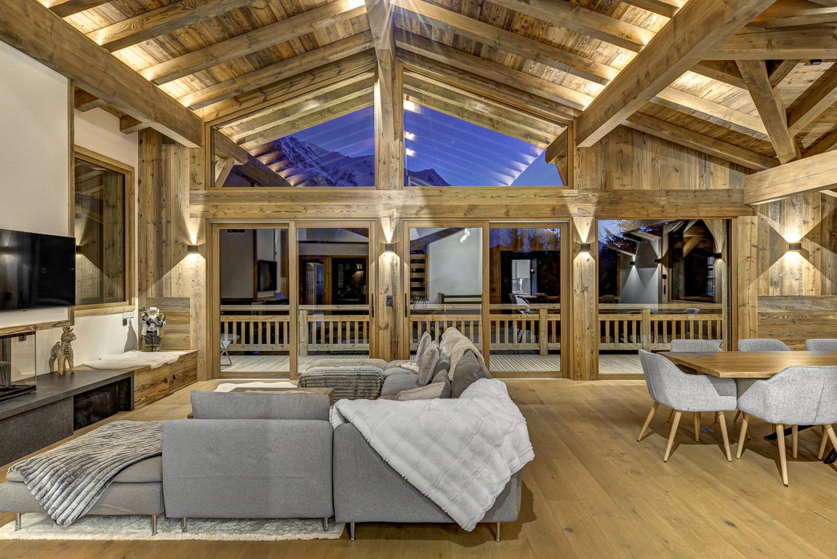 A luxury chalet-style living room with a large gray sectional sofa, dining table with gray chairs, exposed wooden beams, and floor-to-ceiling windows looking out to mountains at dusk.