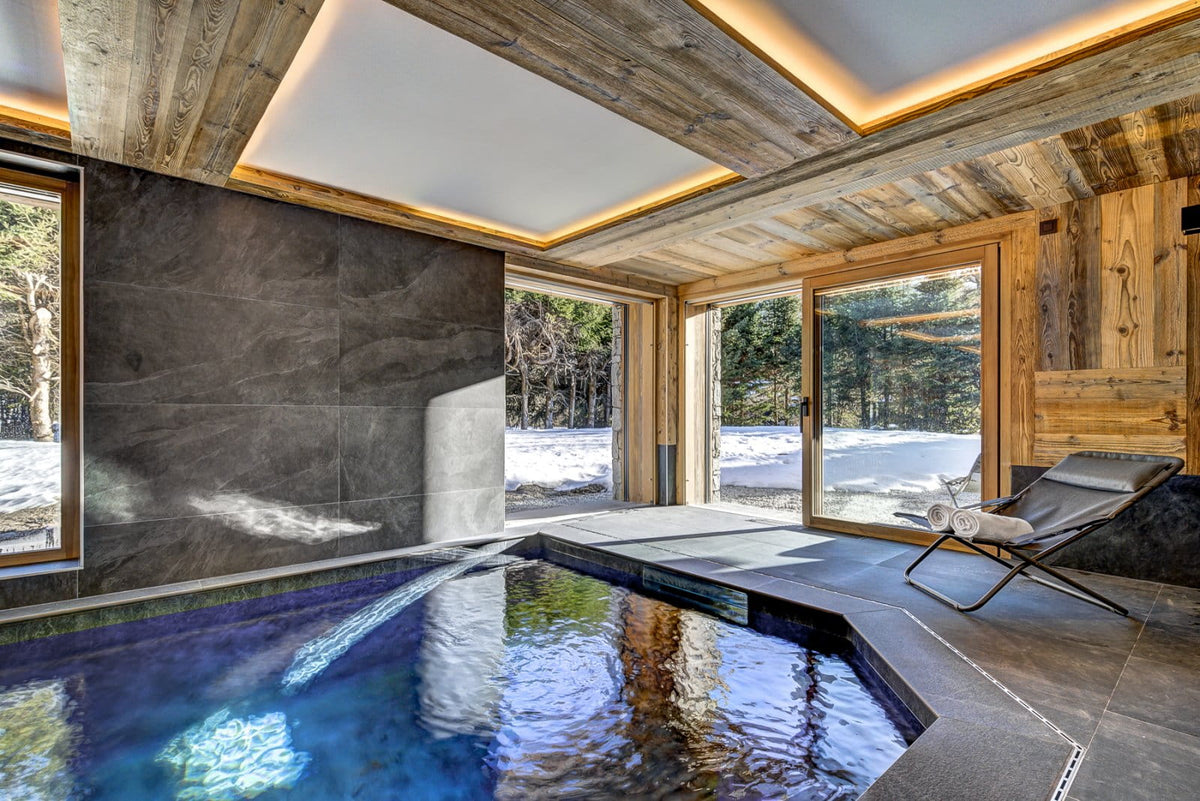 A luxury indoor ski resort pool with a dark stone tiled wall, wooden ceiling beams with warm LED lighting, and large glass doors overlooking a snowy outdoor forest view. A modern lounge chair sits on the deck.