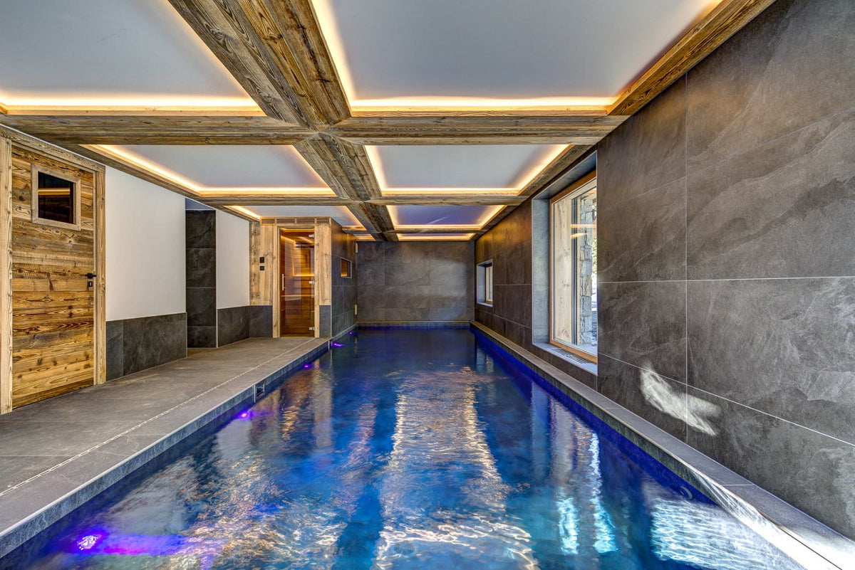 An indoor long pool with blue water, dark stone walls, wood ceiling beams, and warm recessed lighting. A wooden door with a small window is visible on the left, and windows line the right wall.