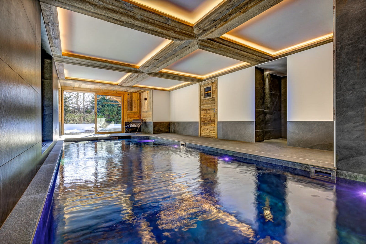 An indoor heated pool in a luxury modern spa or resort, featuring a high coffered ceiling with warm LED lighting, wood paneling, and stone walls.