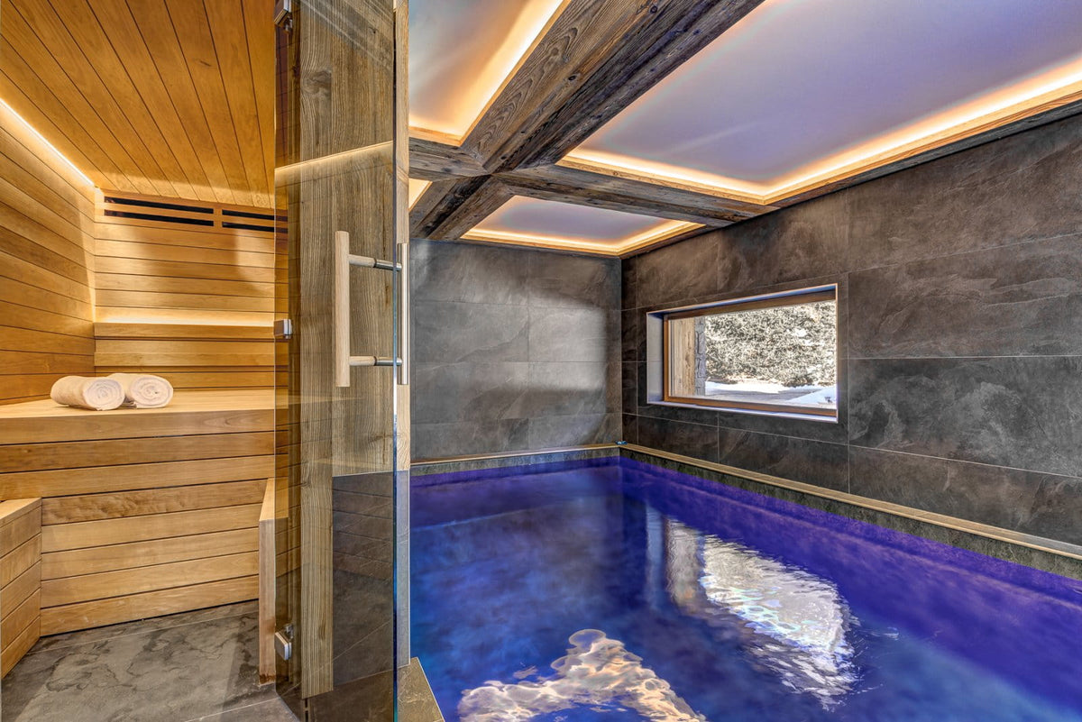 Luxury indoor spa or sauna room with a glass door opening to a wooden sauna and a blue heated pool area with dark stone walls and a window.
