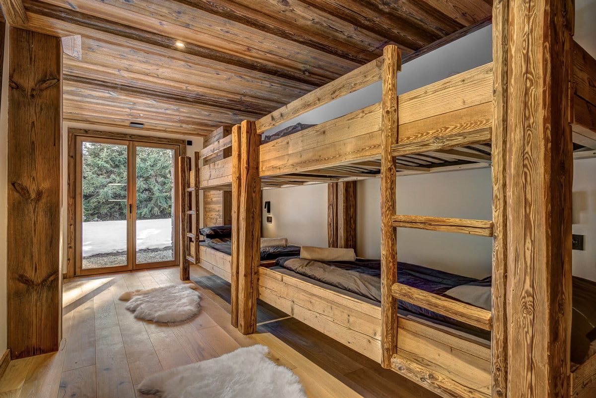 A rustic wooden bunk bed with a ladder, situated in a cozy cabin bedroom with exposed wood ceiling beams, light wood floors, and a large window looking out onto trees.