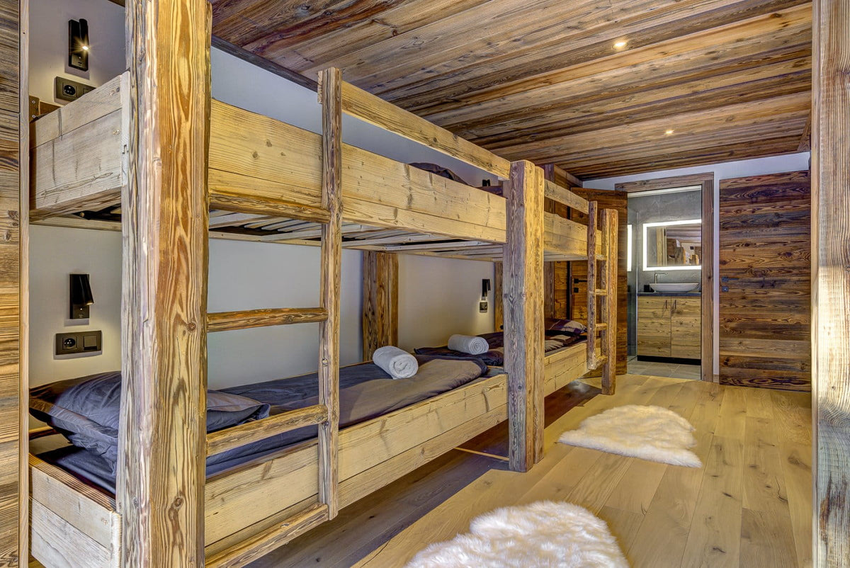 A rustic wooden bunk bed with gray bedding in a cozy, wood-paneled bedroom. White faux-fur rugs are on the light wood floor, and a bathroom is visible in the background.