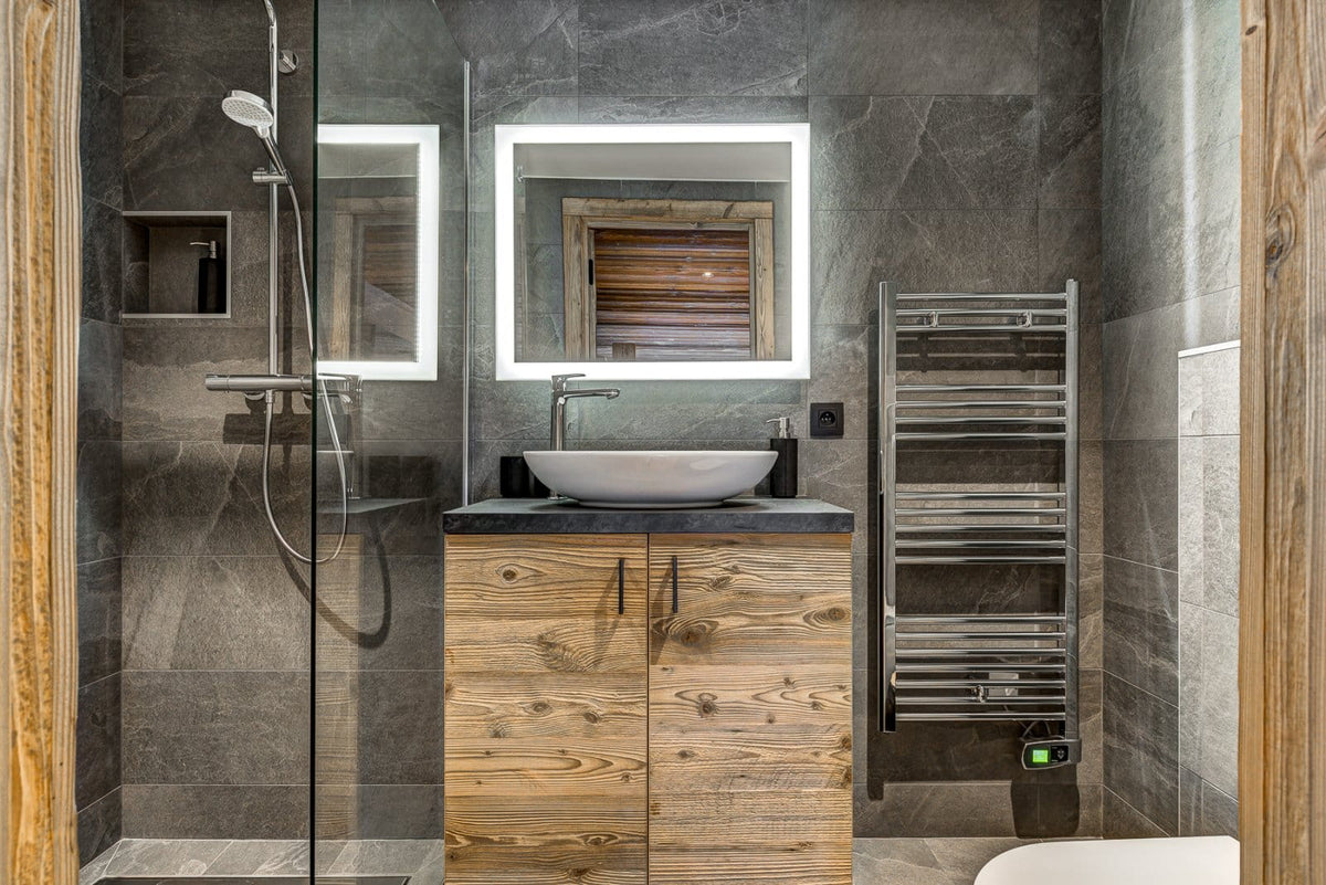 A modern bathroom interior featuring a wood-look vanity cabinet with a white vessel sink, a backlit rectangular mirror, a glass shower enclosure, a heated towel rail, and dark stone tile walls.