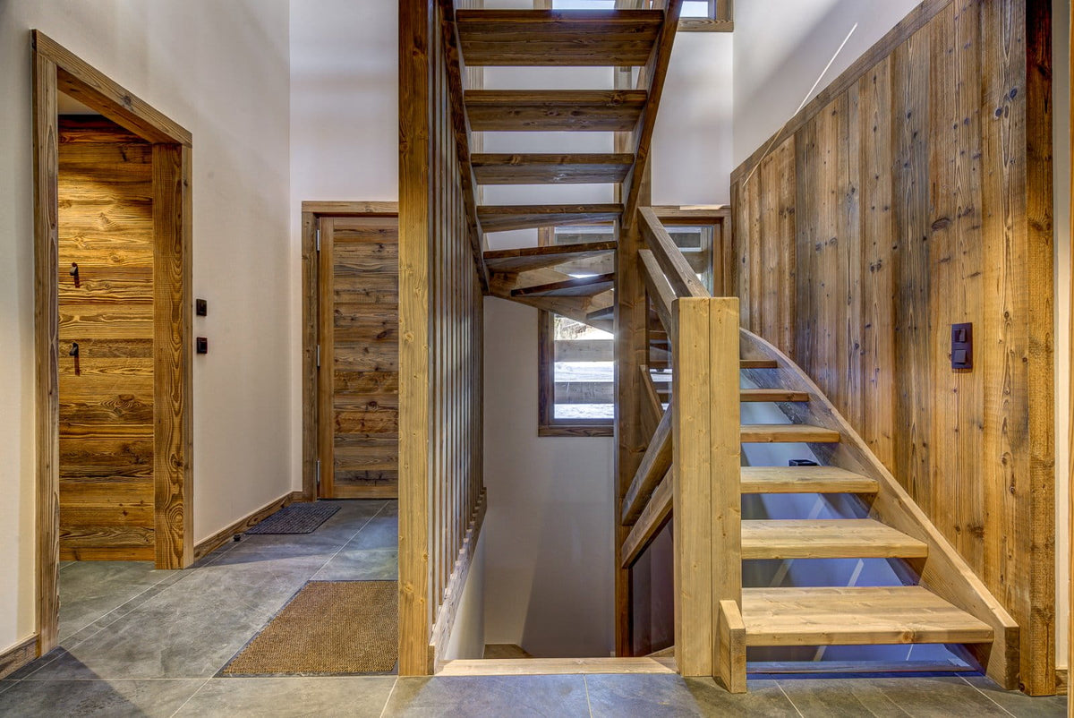 A rustic wooden staircase with light oak steps and a matching wood banister, set against a wall of dark wood paneling in a modern home interior.