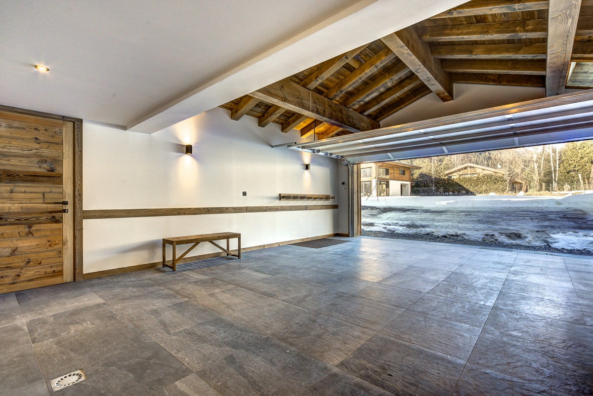 A large, modern garage interior with gray stone floor tiles, white walls, exposed wooden ceiling beams, a wooden bench, and an open overhead door revealing a snowy outdoor landscape.