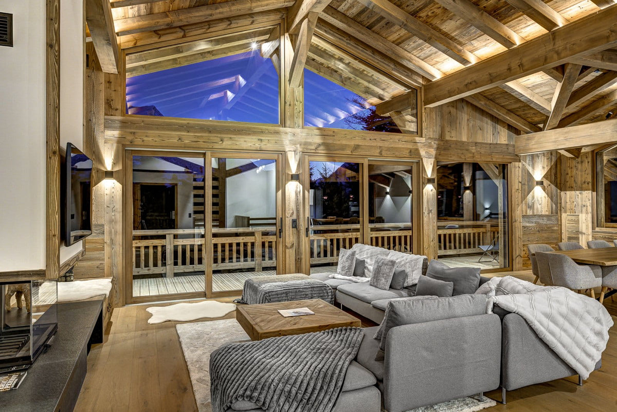 A modern gray sectional sofa in a rustic wooden chalet living room with high ceilings, large windows, and a view of an outdoor deck at night.