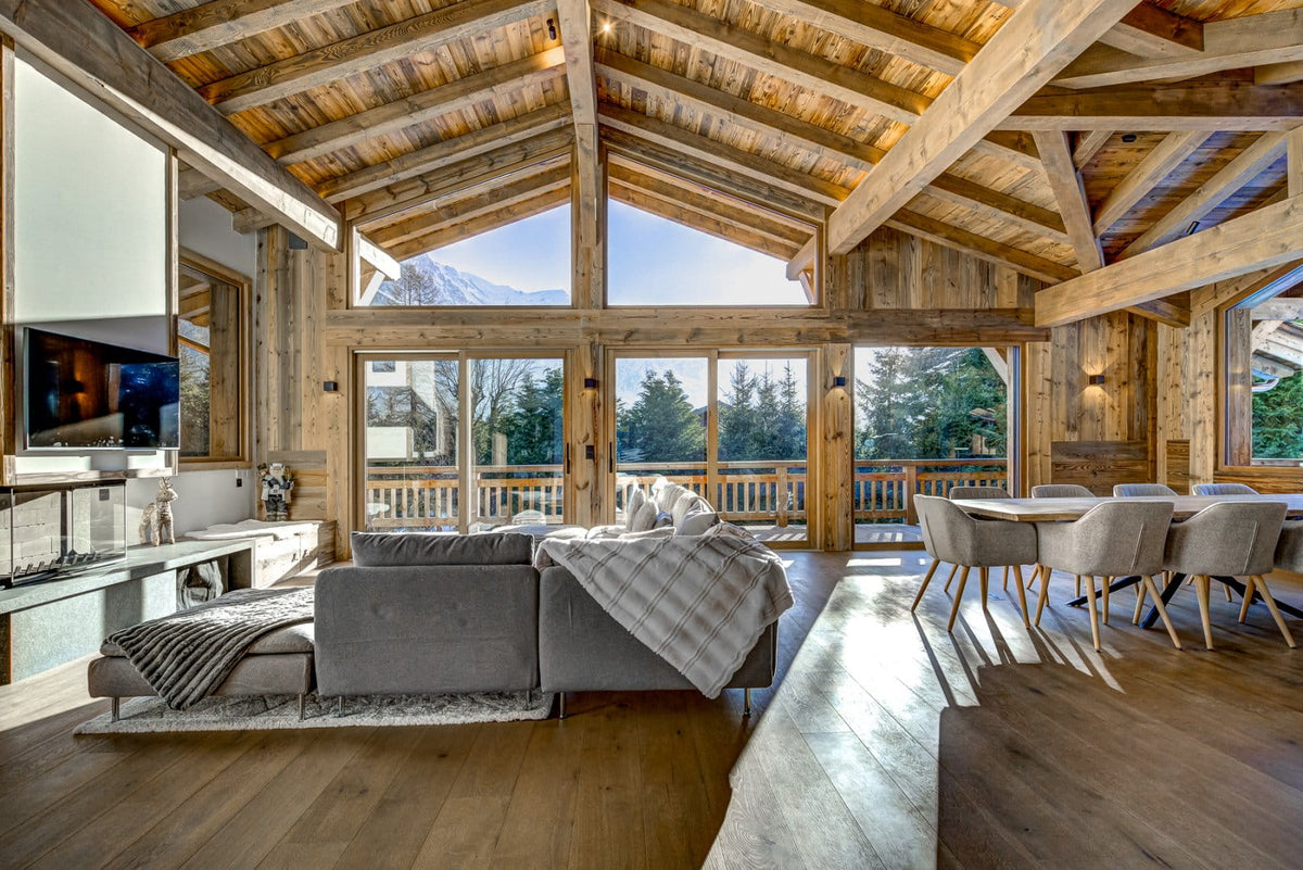 A luxury modern chalet interior with a gray sectional sofa, dining area, and large windows looking out to snowy mountains.