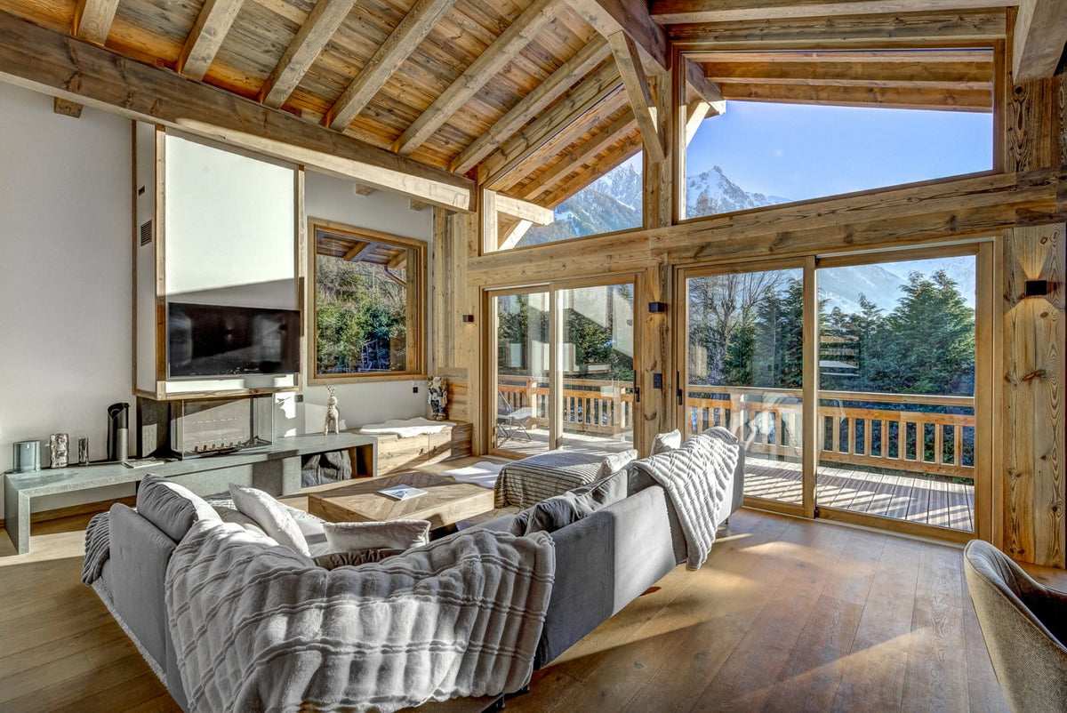 A modern luxury chalet living room featuring a large gray sectional sofa with a textured throw blanket, a wall-mounted flat-screen TV, and massive wood-framed windows looking out to mountains and a balcony.