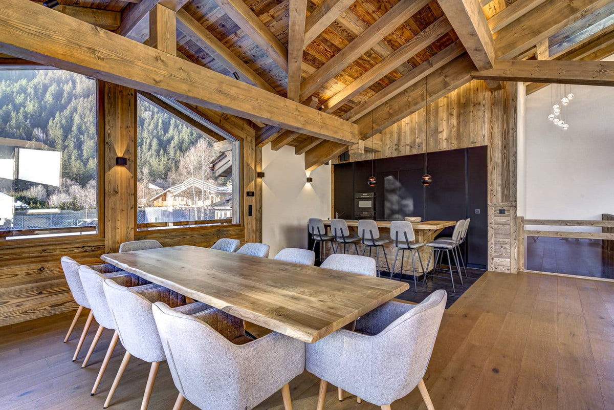 A rustic modern dining room with a long wooden table and light gray upholstered chairs, set in a cabin with exposed wood beams, large windows, and a mountain view.
