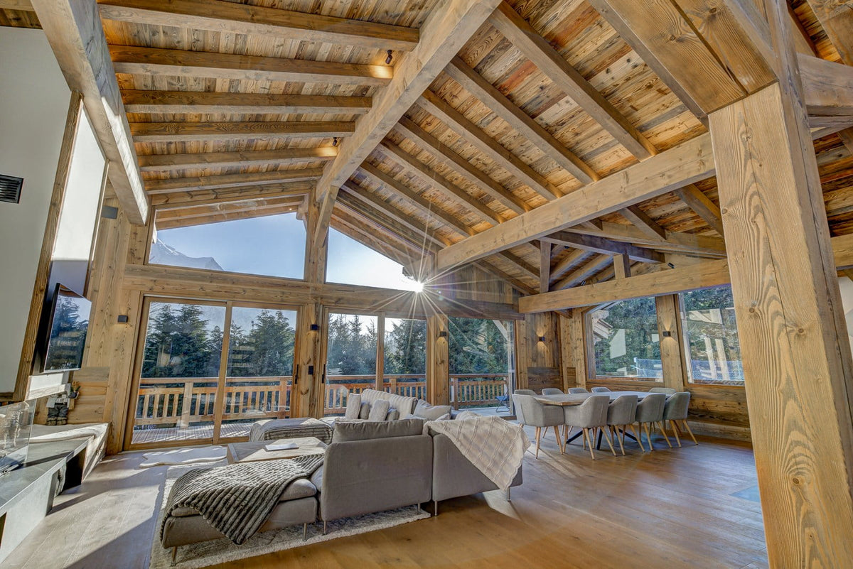 A high-ceilinged rustic lodge-style living room with exposed wooden beams, large floor-to-ceiling windows, a gray sectional sofa, a dining area with gray chairs, and a visible outdoor mountain view.