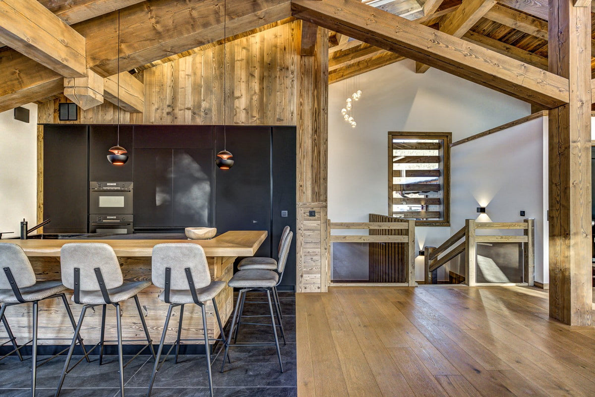 A modern rustic kitchen and dining area with exposed wooden beams, a wooden island, gray upholstered bar stools, a black kitchen cabinetry wall, and a staircase in the background.