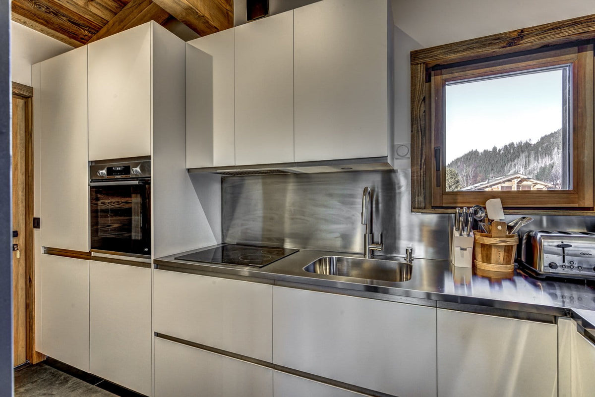 Modern kitchen cabinetry with matte white doors, stainless steel countertops and backsplash, built-in oven, sink, and cookware, set in a rustic interior with exposed wood ceiling beams and a window showing a mountain view.
