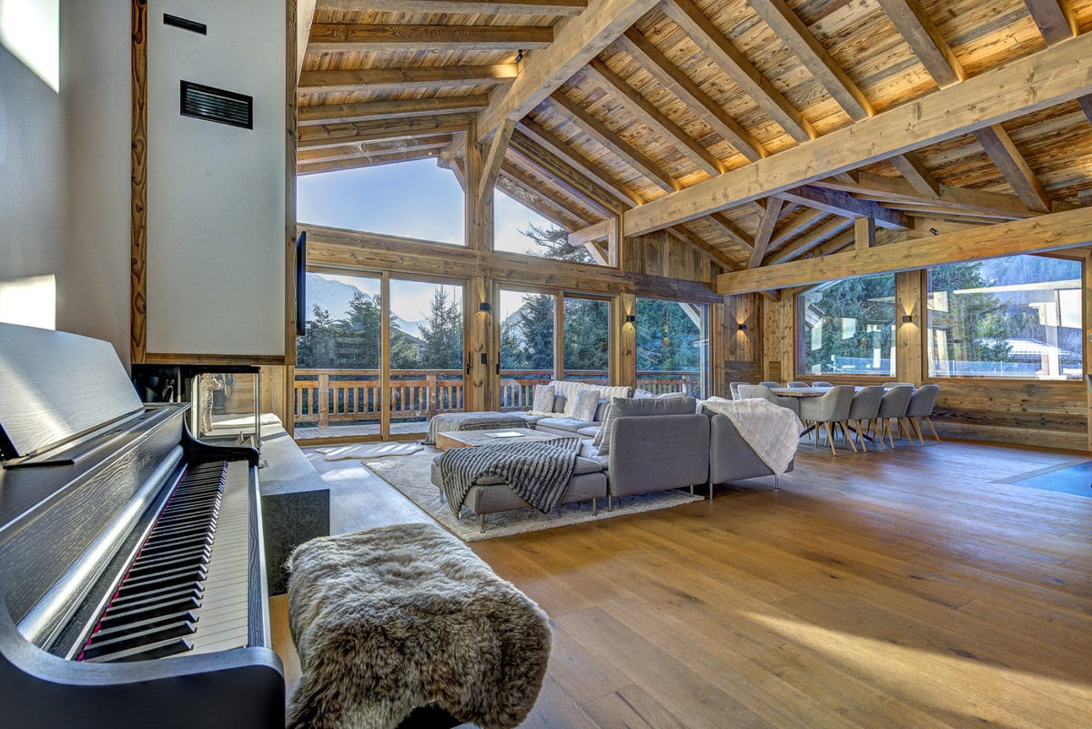 A large, elegant ski chalet living room with high wooden beams, hardwood floors, a gray sofa, and a black digital piano in the foreground.