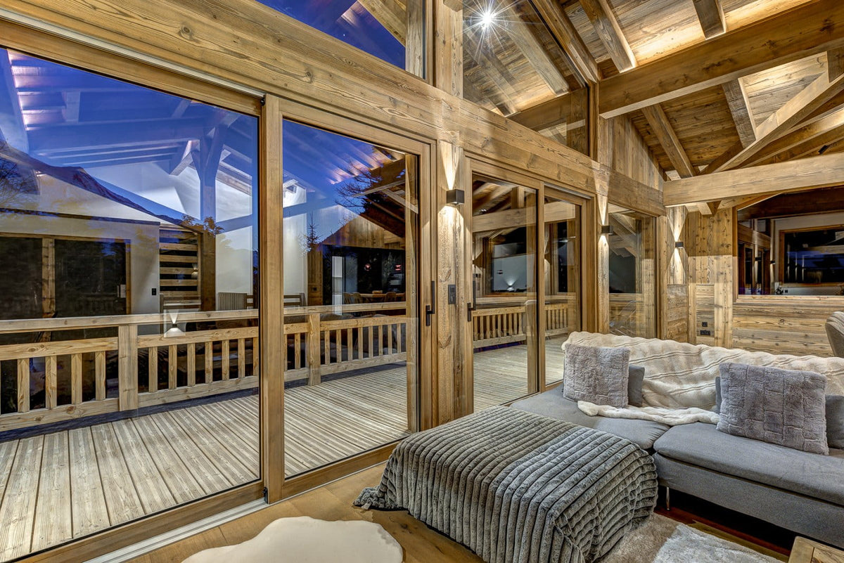 A luxury chalet-style living room with high wooden ceilings, large glass sliding doors opening to a deck at dusk, a gray sectional sofa with textured pillows, and warm ambient lighting.