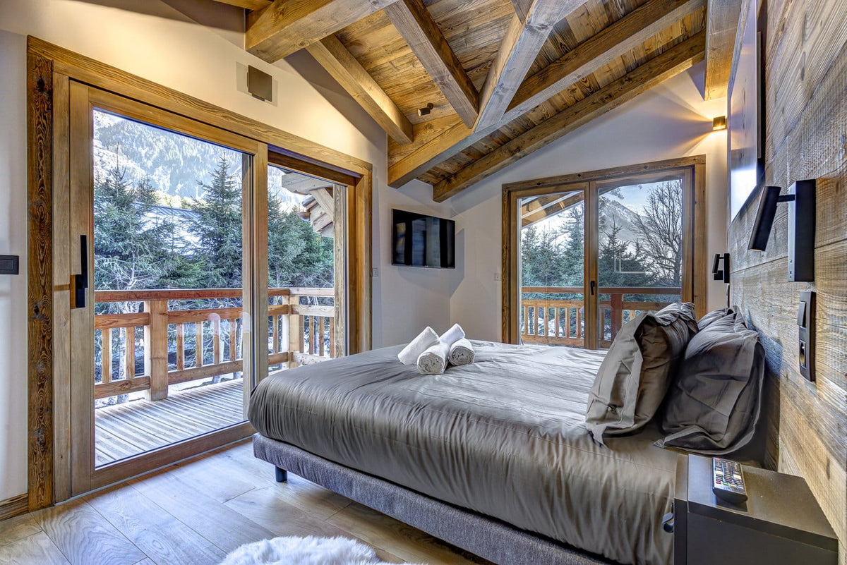 A modern mountain chalet bedroom featuring a large bed with gray bedding, wooden ceiling beams, and floor-to-ceiling windows overlooking a snowy mountain landscape and wooden balcony.