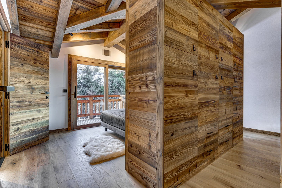 A large wooden wardrobe or closet with rustic handcrafted wood panels, set in a modern chalet-style bedroom with wood floors, a white sheepskin rug, and a balcony visible in the background.