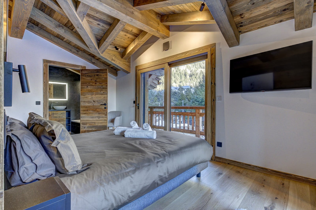 A modern rustic bedroom featuring a large bed with gray bedding, a wooden ceiling with exposed beams, a wall-mounted flat-screen TV, and large windows looking out to a balcony and outdoor scenery.