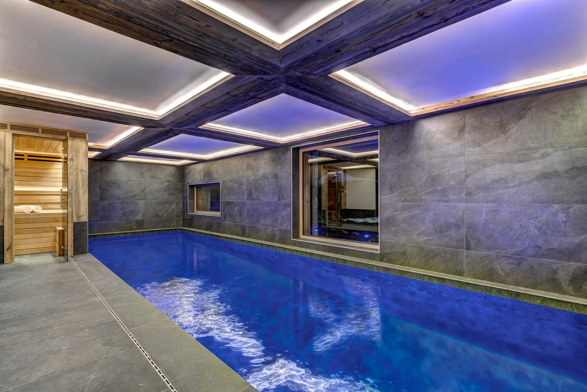 An indoor swimming pool with bright blue water, dark stone walls, and a wooden sauna visible on the left, set under a modern ceiling with recessed LED lighting.
