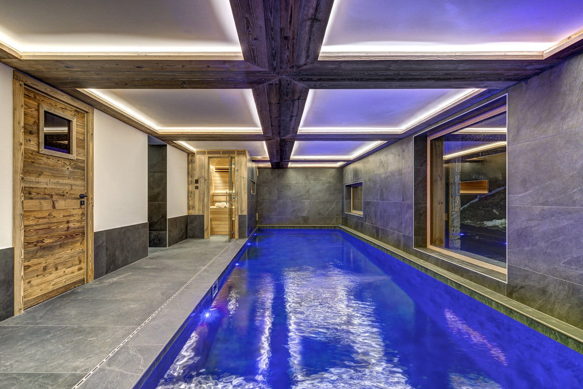 An indoor long pool with vivid blue water, set in a modern mountain-style room with gray stone walls, dark wood ceiling beams, and recessed LED lighting.