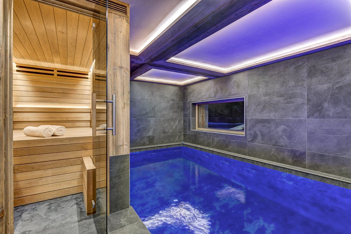 An indoor spa facility featuring a wooden sauna with rolled towels and a rectangular indoor pool with vivid blue water, set against dark gray stone walls and ceiling with purple ambient lighting.