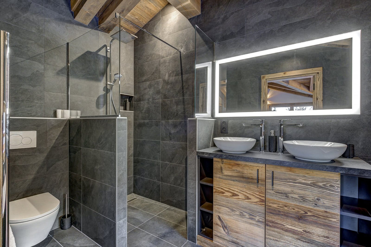 Modern bathroom interior with dark gray stone tile walls and floor, a glass shower enclosure, a white toilet, a double vessel sink vanity with wood cabinets, and a large backlit mirror.