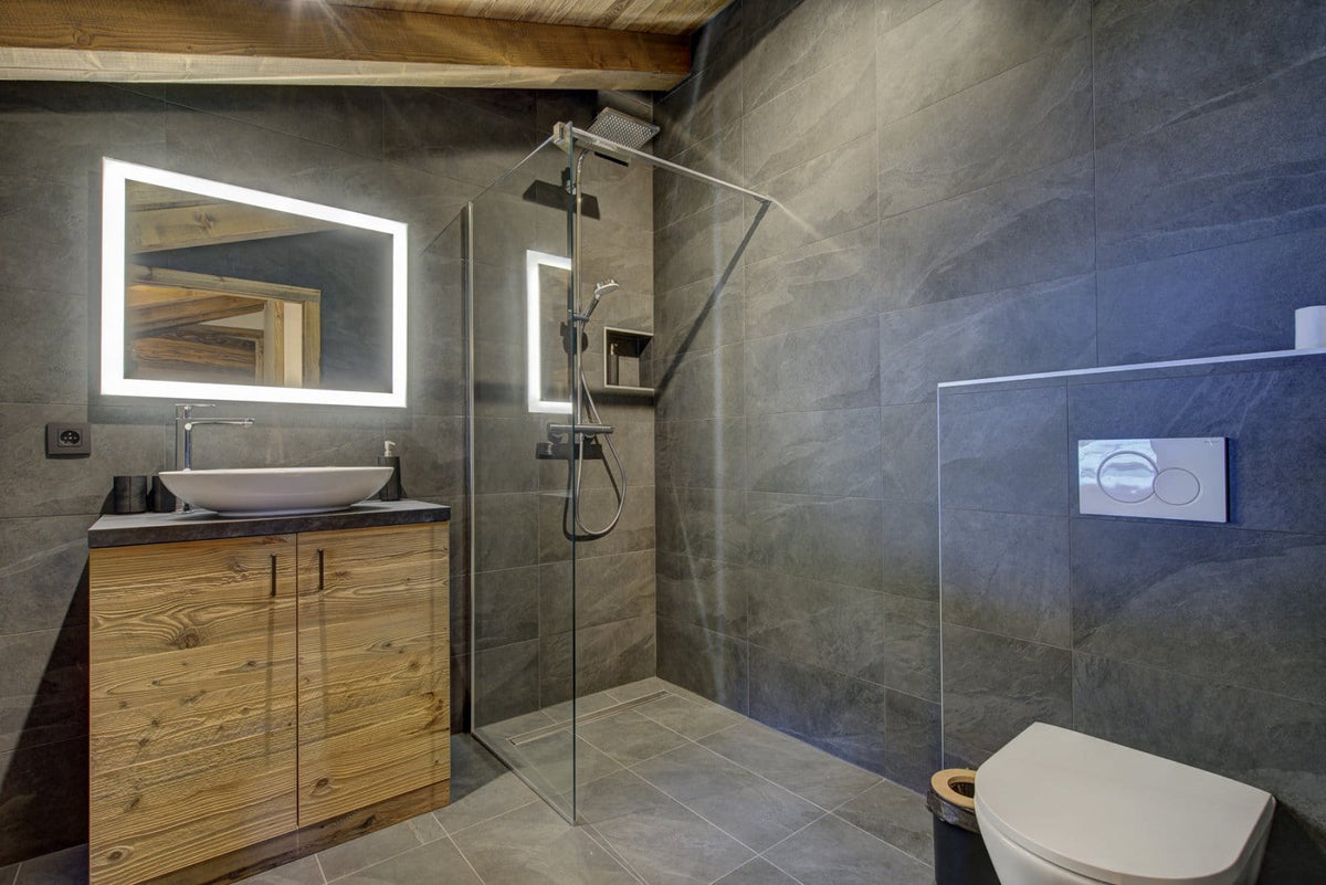 Modern bathroom interior with a wood vanity, illuminated mirror, glass shower enclosure, dark stone walls, and a white toilet.