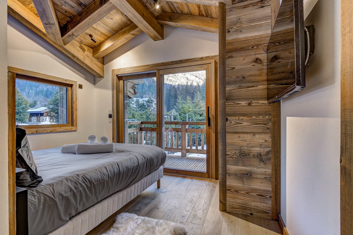 A modern bedroom interior with a bed dressed in gray bedding, large wood-framed windows overlooking a mountain view, exposed wooden ceiling beams, and a wood-paneled wall with a mounted TV.