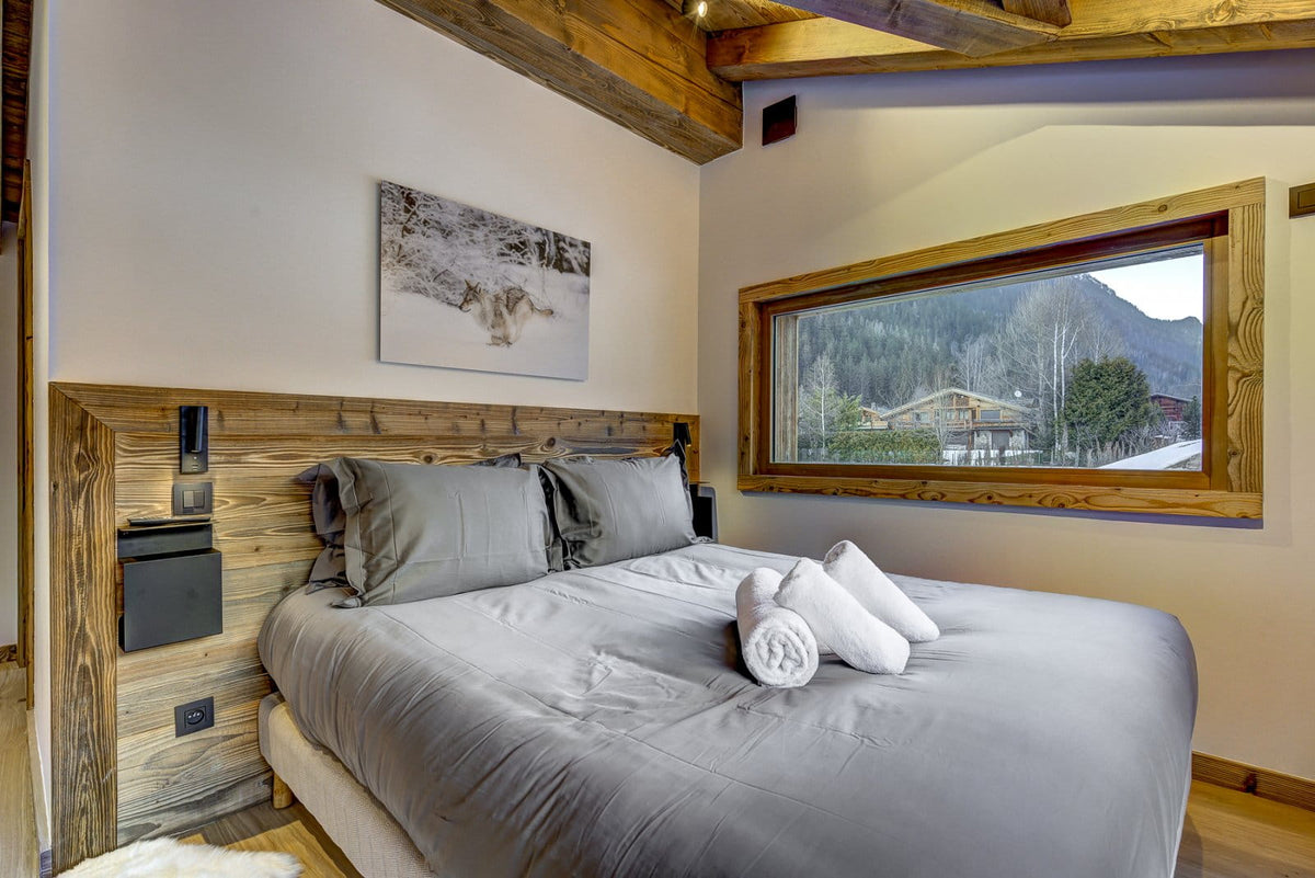 A modern bedroom featuring a large bed with gray bedding, a rustic wooden headboard and ceiling beams, and a large window showing a mountain landscape outside.
