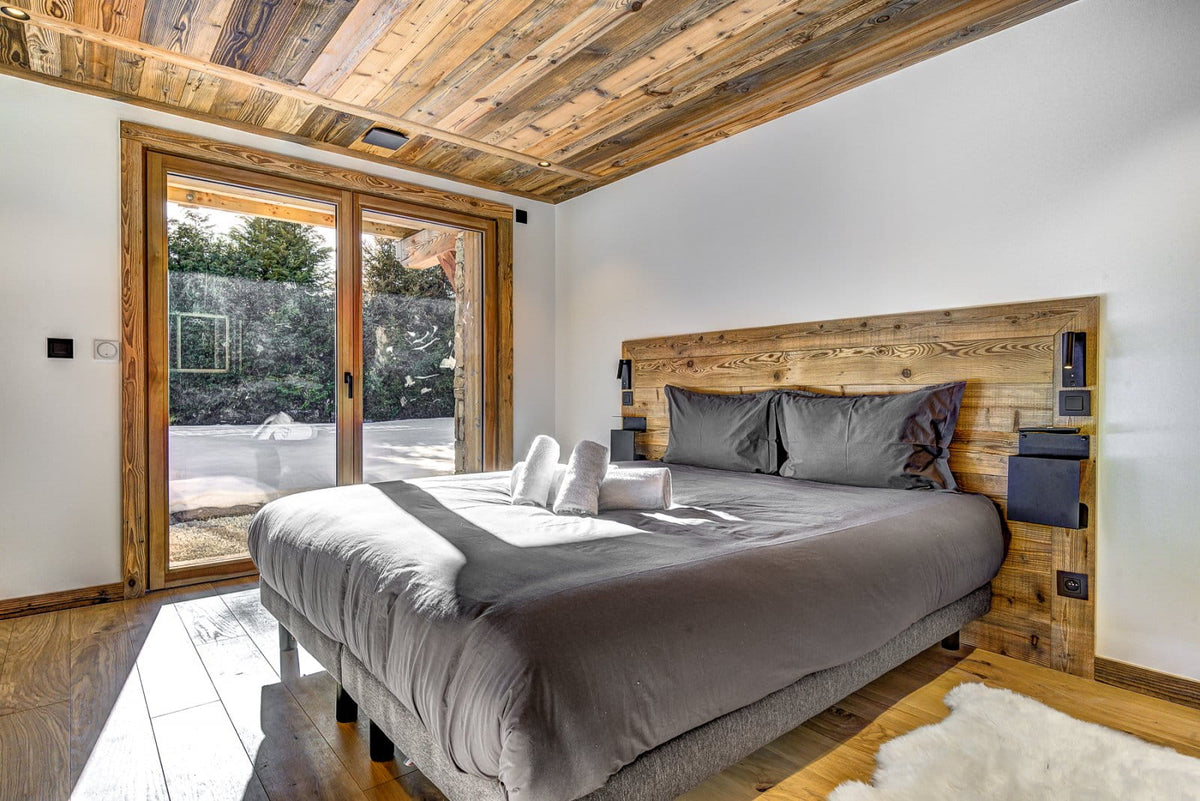 A modern bedroom featuring a large bed with gray bedding and a rustic wooden headboard, set against white walls with a wood-paneled ceiling and a glass door leading to an outdoor patio.