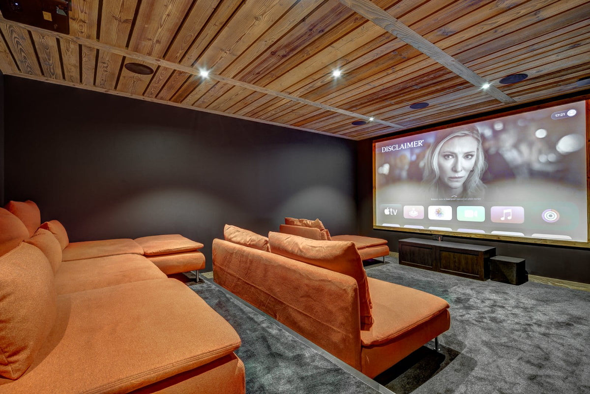 A home theater setup featuring plush orange sectional sofas arranged in rows facing a large projection screen on a dark wall, with a wood-paneled ceiling and recessed lighting.