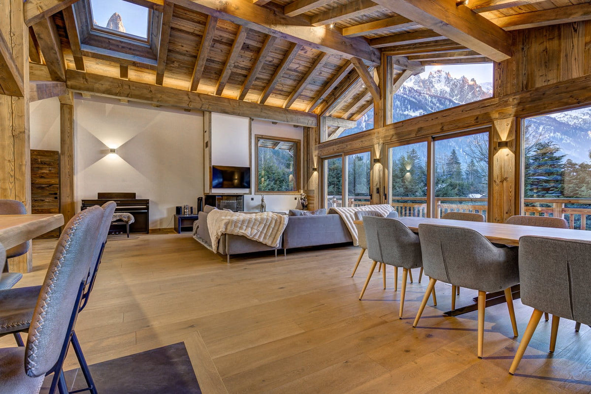 A luxury chalet-style living room with a high wood-beamed ceiling, large windows showing a mountain view, a gray sofa, and dining chairs around a wooden table.