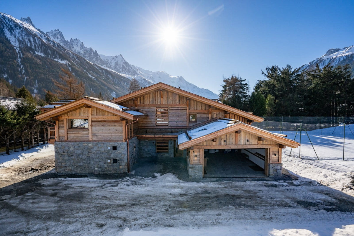A modern wooden chalet-style mountain house with stone foundations and snow-covered roofs, set against a backdrop of snowy peaks and a bright sun in a clear blue sky.