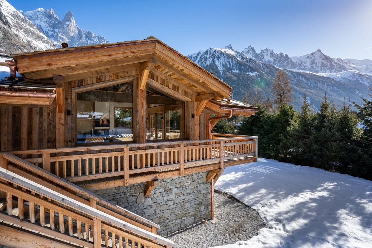 A rustic wooden chalet with stone foundations and large glass windows, set in a snowy mountain landscape with snow-covered peaks under a clear blue sky.