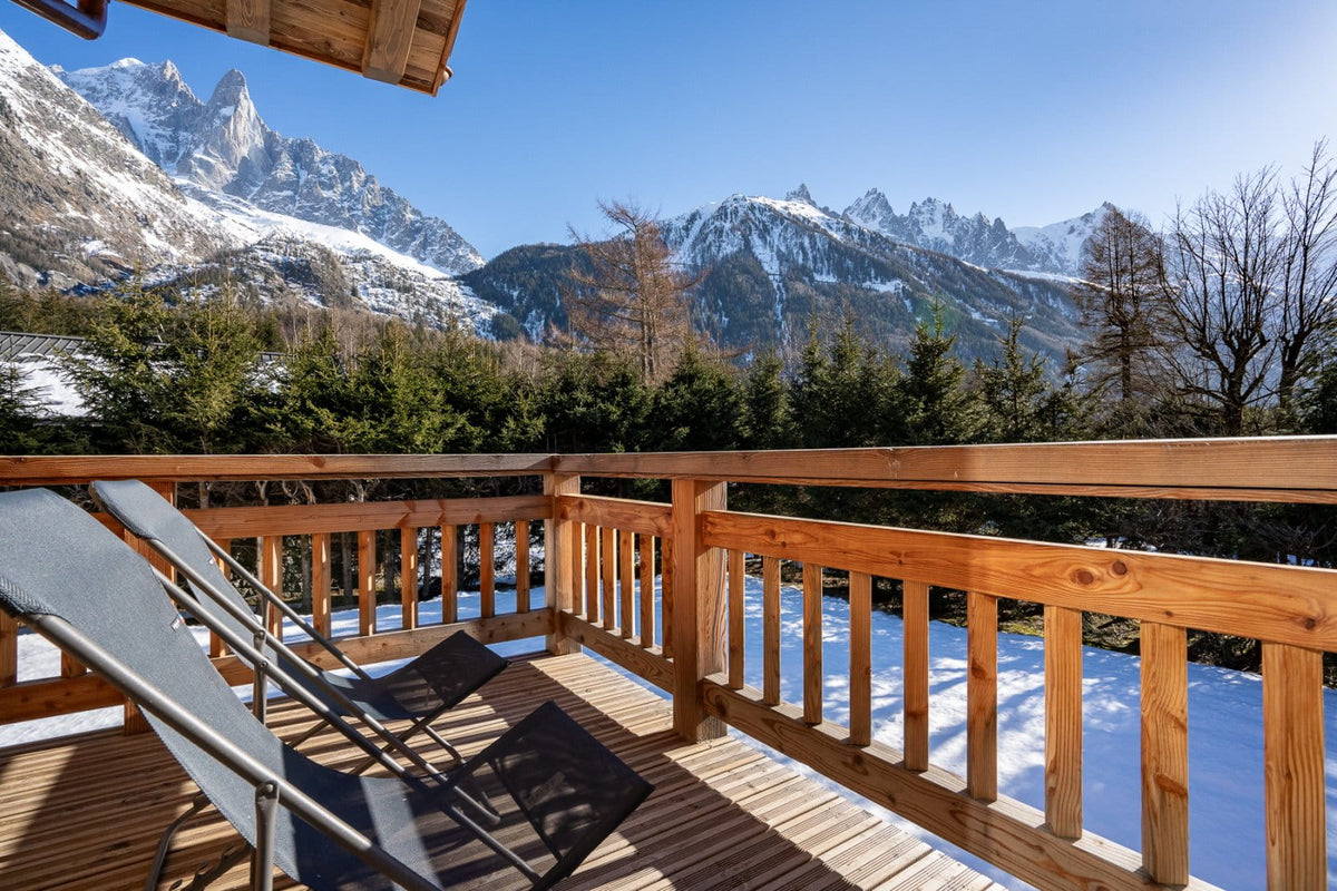 A wooden balcony overlooking a snowy mountain landscape under a clear blue sky, with two dark lounge chairs on the deck.