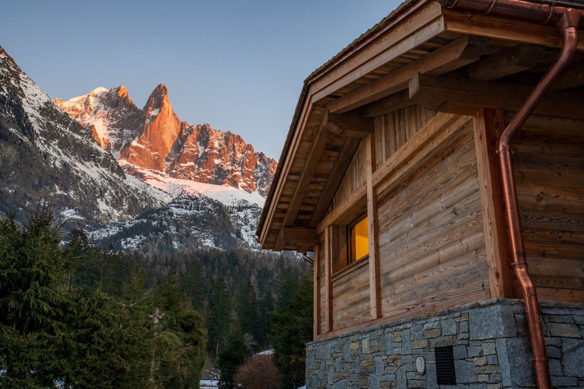 A rustic wooden chalet with a stone base and copper downspout, set against a dramatic mountain landscape with snow-capped peaks at sunrise.